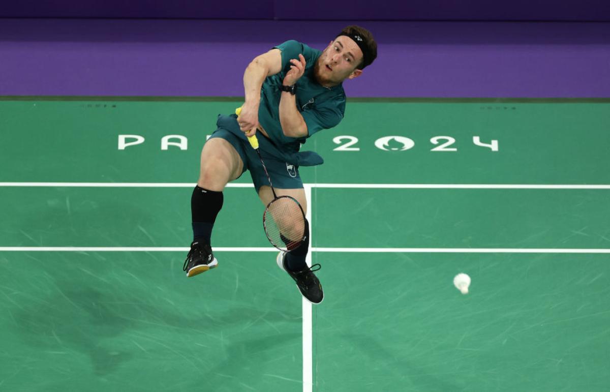 A male Para badminton player is jumping while he smashes a shuttlecock on a green indoor court